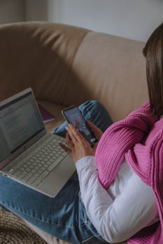 Home Woman sitting on a sofa multitasking with a smartphone and a laptop indoors.
